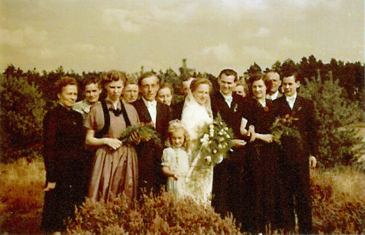 Eine formell gekleidete Hochzeitsgesellschaft versammelt sich auf einer Wiese vor dem Hintergrund von Bäumen und bewölktem Himmel.