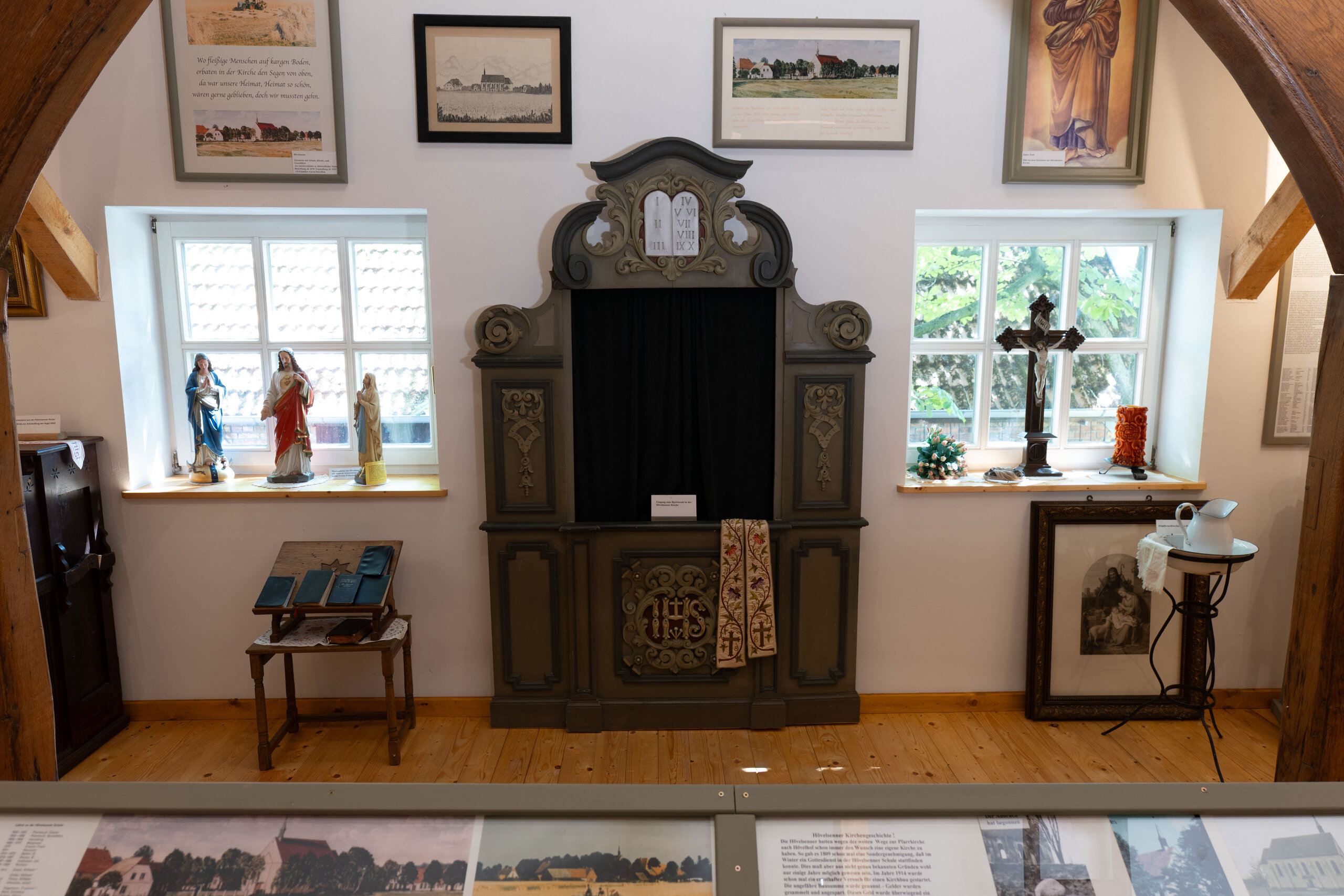 Ein hölzerner Beichtstuhl mit schwarzem Vorhang und goldenen Akzenten, flankiert von zwei Fenstern, ist umgeben von religiösen Artefakten und gerahmten Bildern in einer musealen Umgebung.