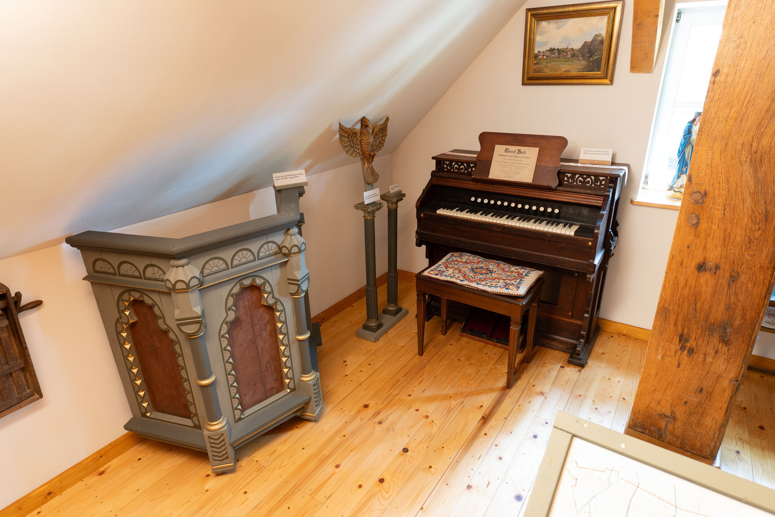 Ein Harmonium mit weißer Tastatur und eine Holzbank mit rotem Kissen stehen in einem Raum mit schräger Decke, Holzbalken und einem Landschaftsgemälde.