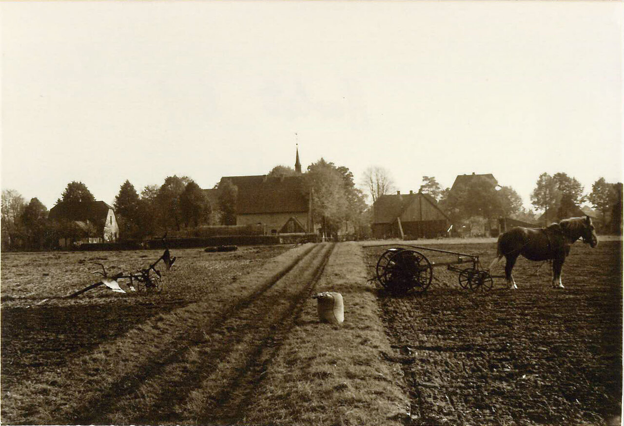 Eine Schwarz-Weiß-Fotografie zeigt eine ländliche Szene mit einem pferdegezogenen Pflug, einem Pferd und einer Kirche in der Ferne, während im Vordergrund ein Feld und Bäume zu sehen sind.