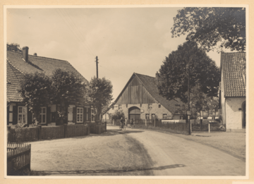 Eine Schwarz-Weiß-Fotografie zeigt eine malerische Dorfszene mit einer unbefestigten Straße, Holzhäusern, einer Scheune und einer Person auf einem Fahrrad.
