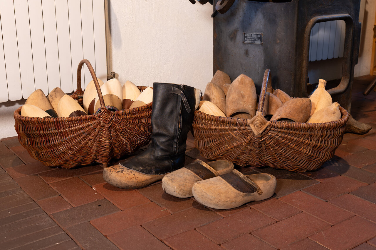 Eine rustikale Szene mit einem schwarzen Stiefel, Holzschuhen und einem mit Holzschuhen gefüllten Weidenkorb vor einer weißen Wand und einem roten Backsteinboden.