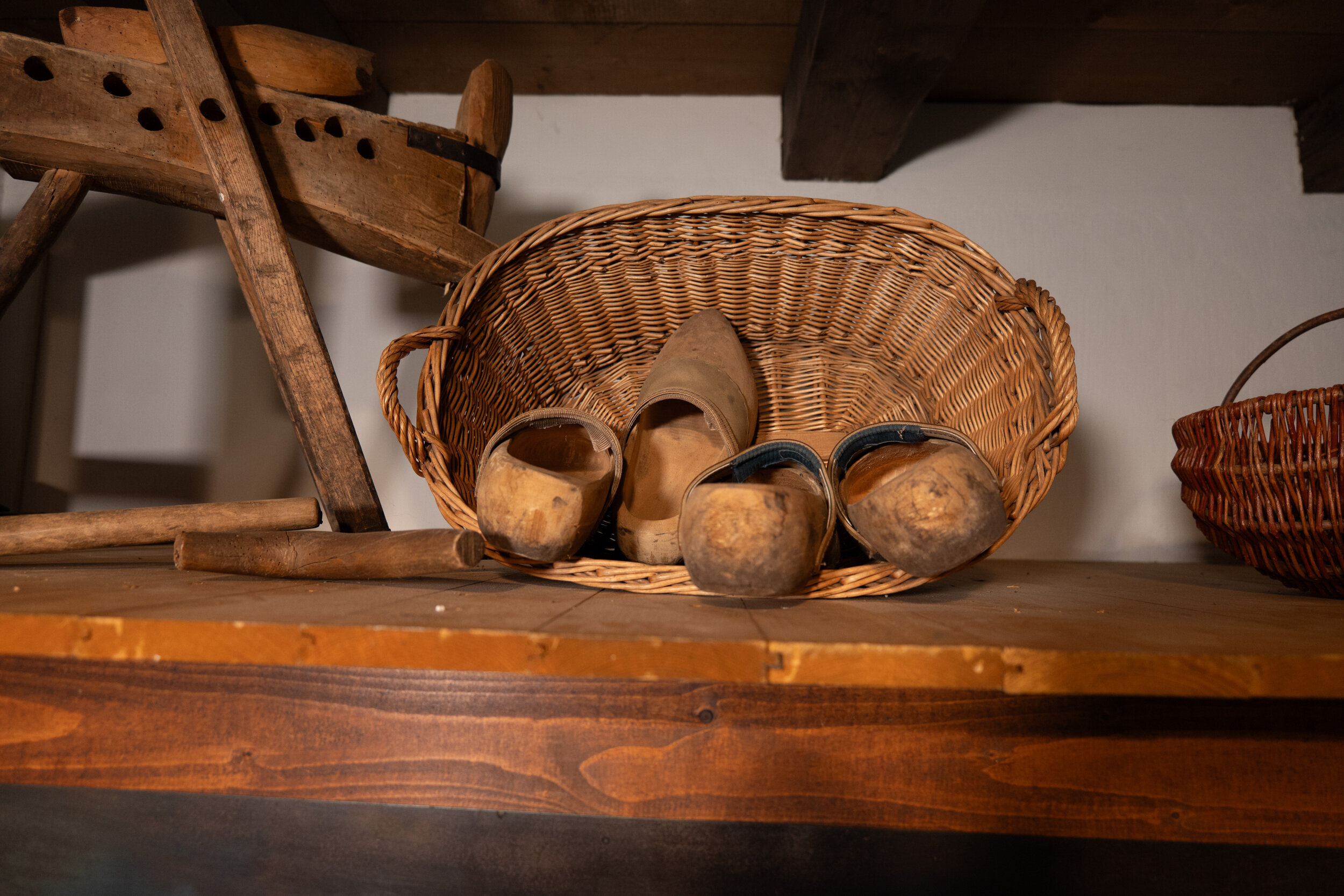 Ein Weidenkorb enthält zwei Paar abgetragene Holzschuhe, daneben stehen ein Holzstuhl und ein roter Korb auf einem Holzregal.