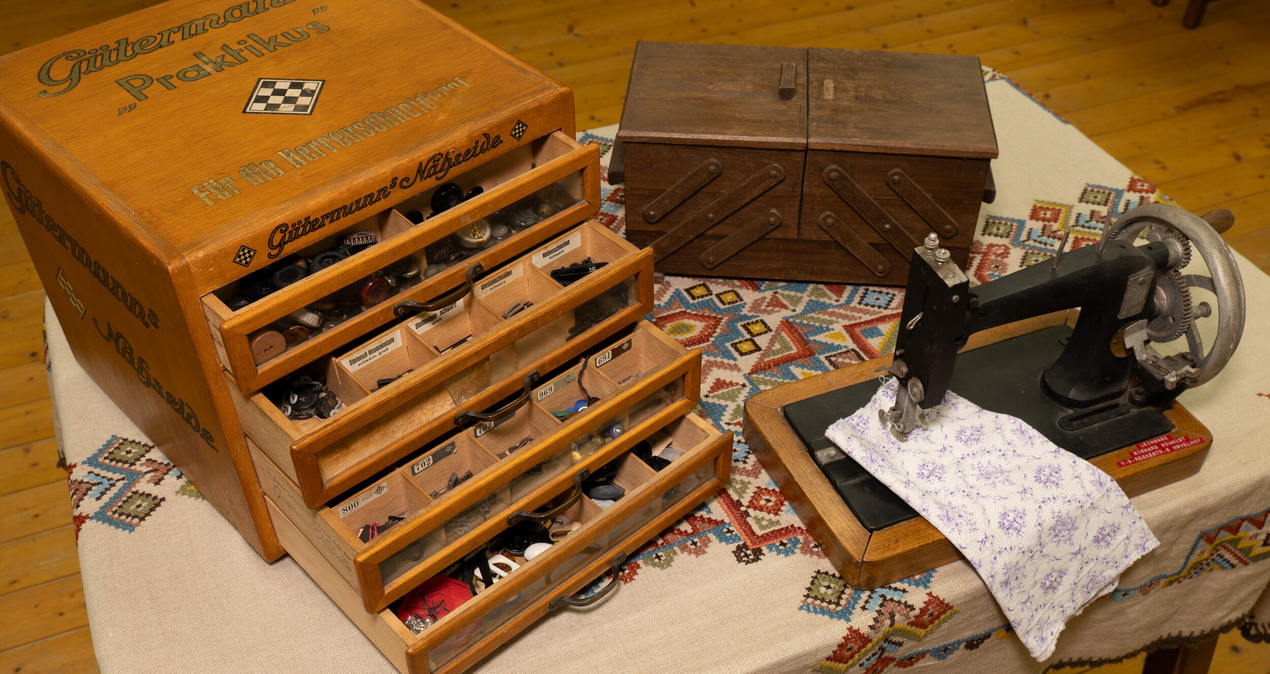 Ein Holzkasten mit der Aufschrift "Gittermann's Praktikus" enthält verschiedene Nähwerkzeuge, dazu eine hölzerne Nähmaschine und eine hölzerne Kommode.