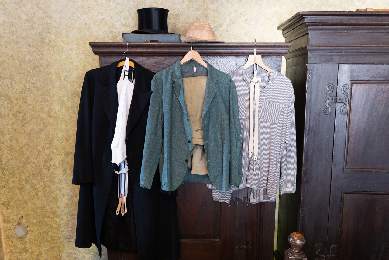 Ein hölzerner Schrank auf dem ein Zylinder liegt. An dem Schrank hängt ein schwarzer Frack mit einem weißen Hemd sowie einer grünen Jacke und ein graues Baumwollhemd sowie eine braune Lederhose.