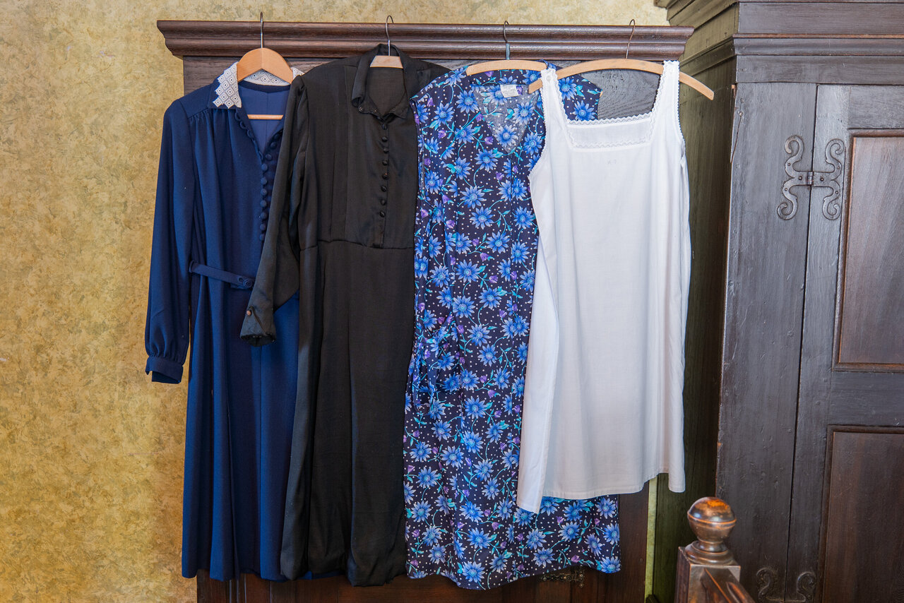 An einem hölzernen Schrank hängen ein blaues und ein schwarzes, langes Kleid, eine geblümte Küchenschürze und ein weißes, langes Unterhemd.