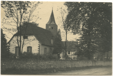 Eine Schwarz-Weiß-Fotografie der evangelischen Kirche in Haustenbeck mit Kirchturm und Uhrenturm, umgeben von Bäumen und einer Steinmauer, wird von einem Baum im Vordergrund eingerahmt.