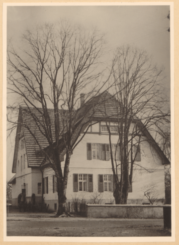 Ein zweistöckiges weißes Haus mit rotem Dach und schwarzen Fensterläden steht auf einer Schwarzweißfotografie, umgeben von kahlen Bäumen und einer Steinmauer.