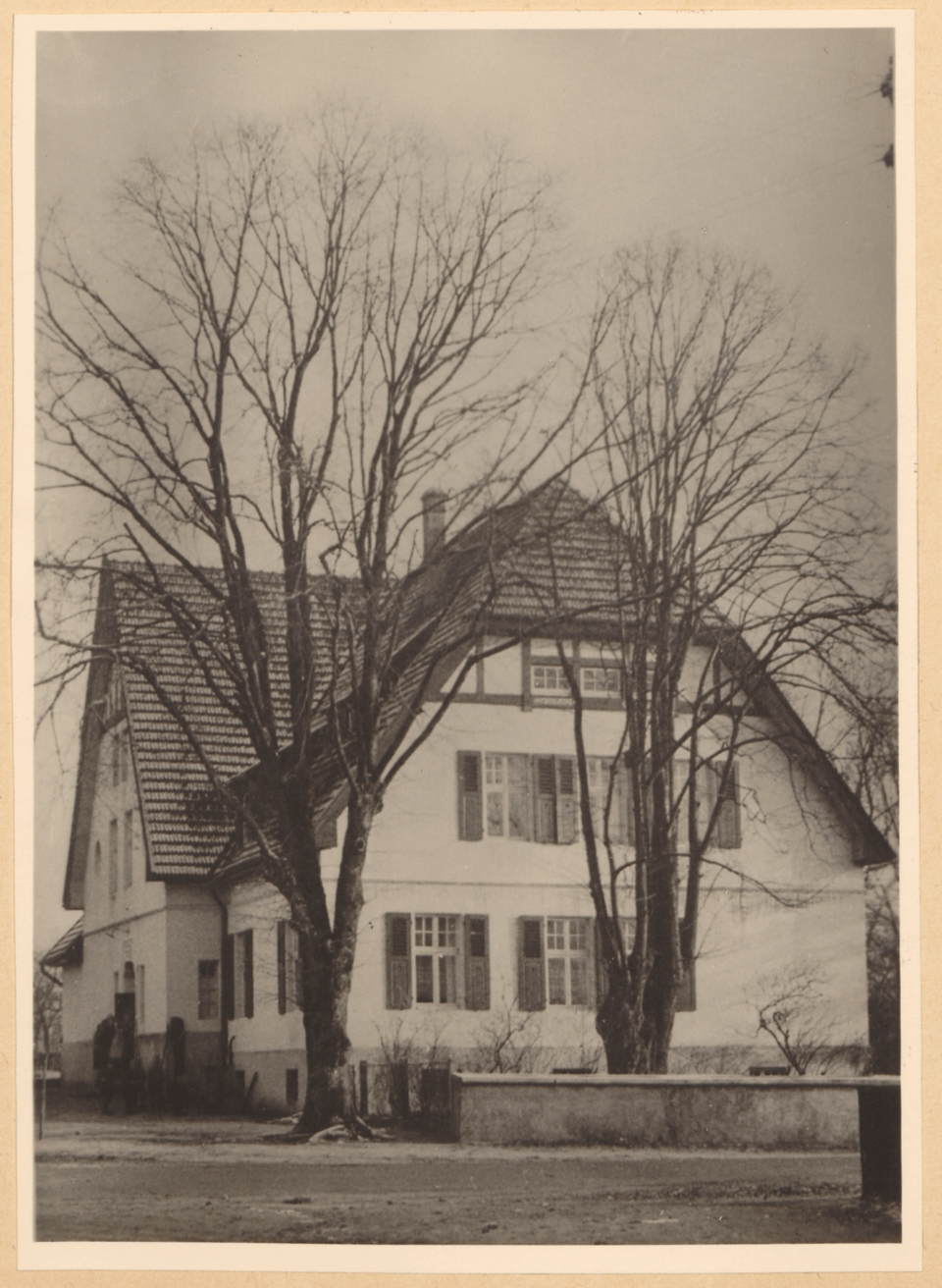 Ein zweistöckiges weißes Haus mit rotem Dach und schwarzen Fensterläden steht auf einer Schwarzweißfotografie, umgeben von kahlen Bäumen und einer Steinmauer.
