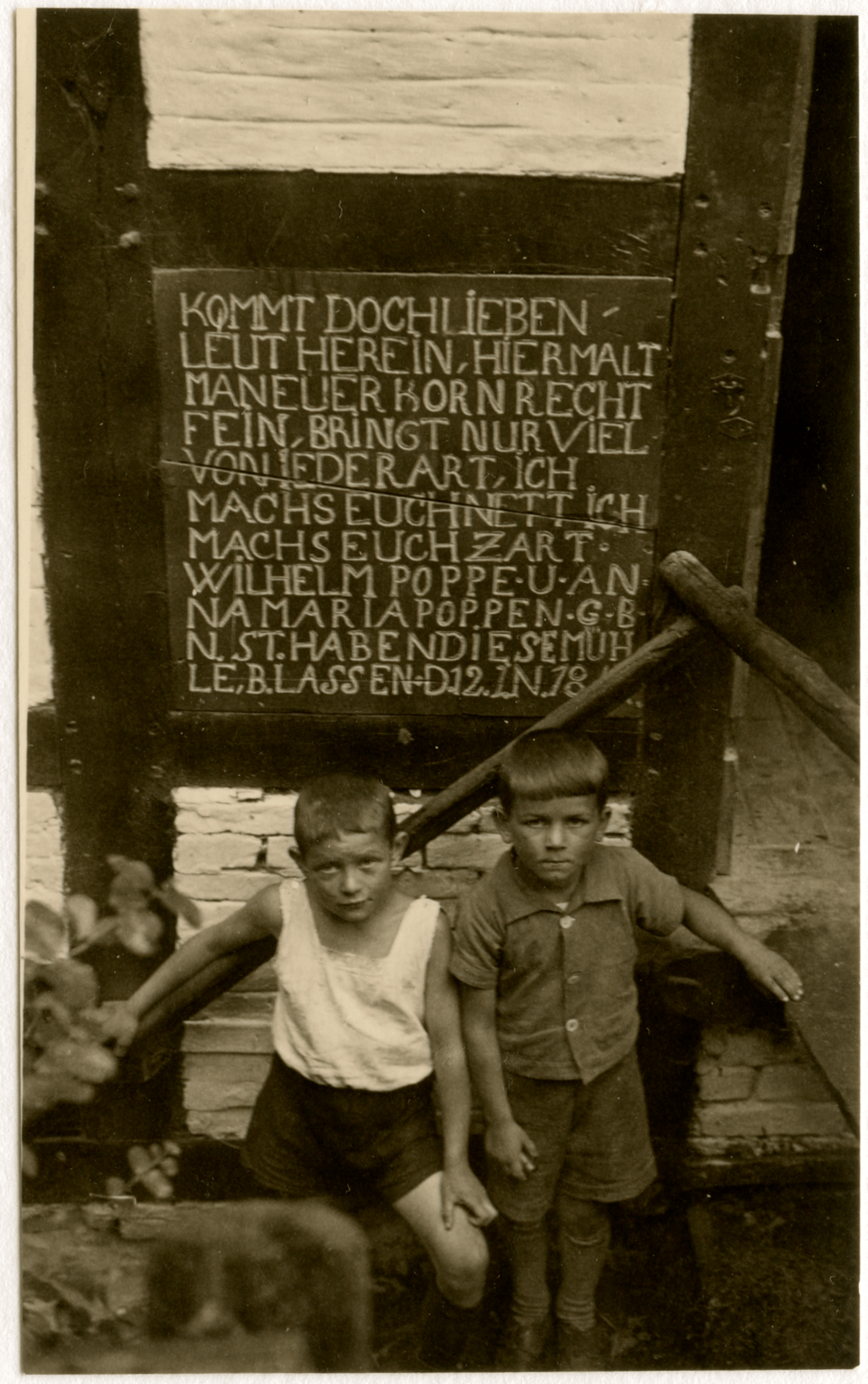 Zwei Jungen in weißen Hemden und kurzen Hosen stehen auf einer Holztreppe,  über ihnen eine Inschriftentafel links neben der Mühlentür in Haustenbeck.