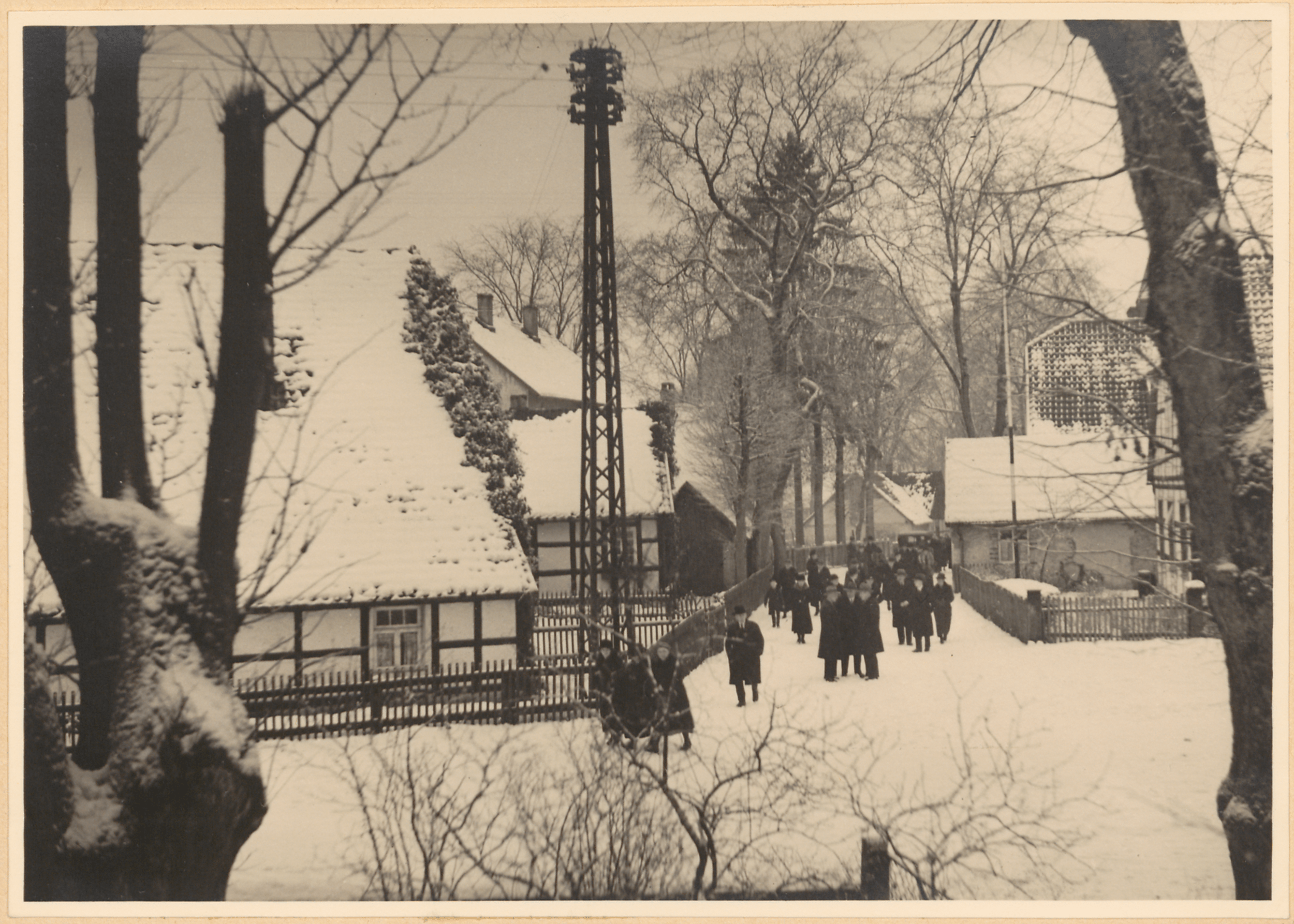 Auf einer verschneiten Dorfstraße ist eine Schwarz-Weiß-Fotografie zu sehen, die eine Gruppe von Spaziergängern mit einem hohen Turm und schneebedeckten Bäumen im Hintergrund zeigt. Die Menschen gehen zur Kirche.