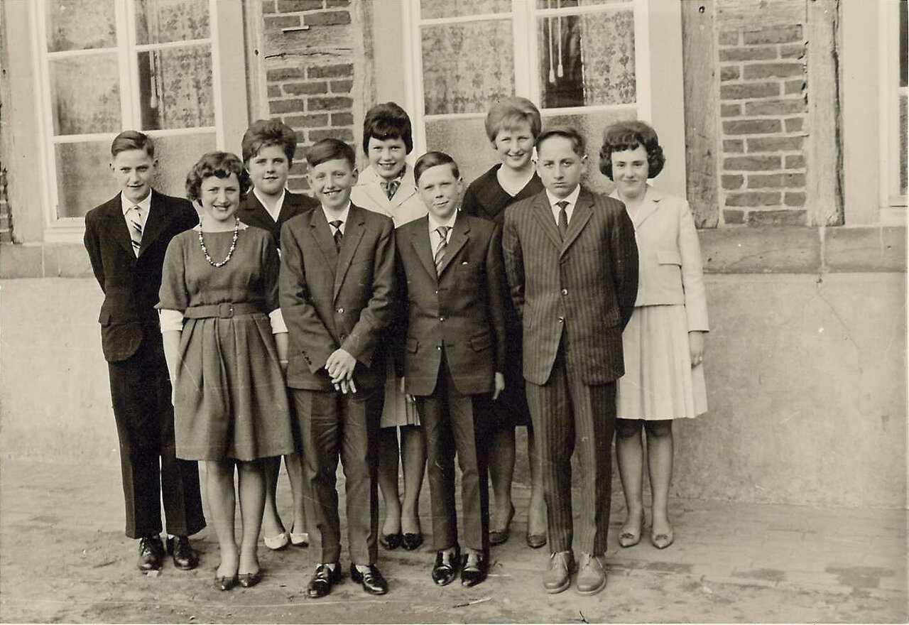 Neun formell gekleidete junge Personen posieren vor einem Backsteingebäude, darunter zwei Mädchen und sieben Jungen, in einer Schwarz-Weiß-Aufnahme. Es handelt sich um die Schulentlassung im April 1962.