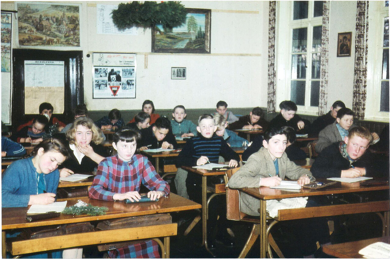 Ein Klassenzimmer mit Schülern, die an hölzernen Schreibtischen sitzen und konzentriert arbeiten, mit einem Gemälde an der Wand und einer Pinnwand darüber.