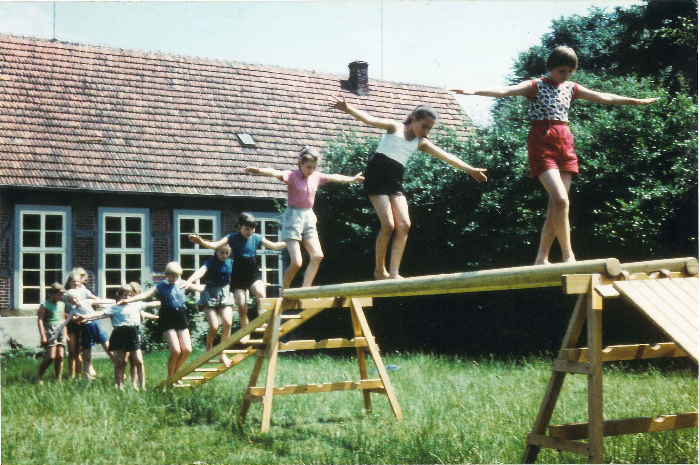 Kinder in farbenfroher Kleidung spielen auf einem hölzernen Hindernisparcours in einer üppigen grünen Wiese, mit einem rotgedeckten Gebäude im Hintergrund.