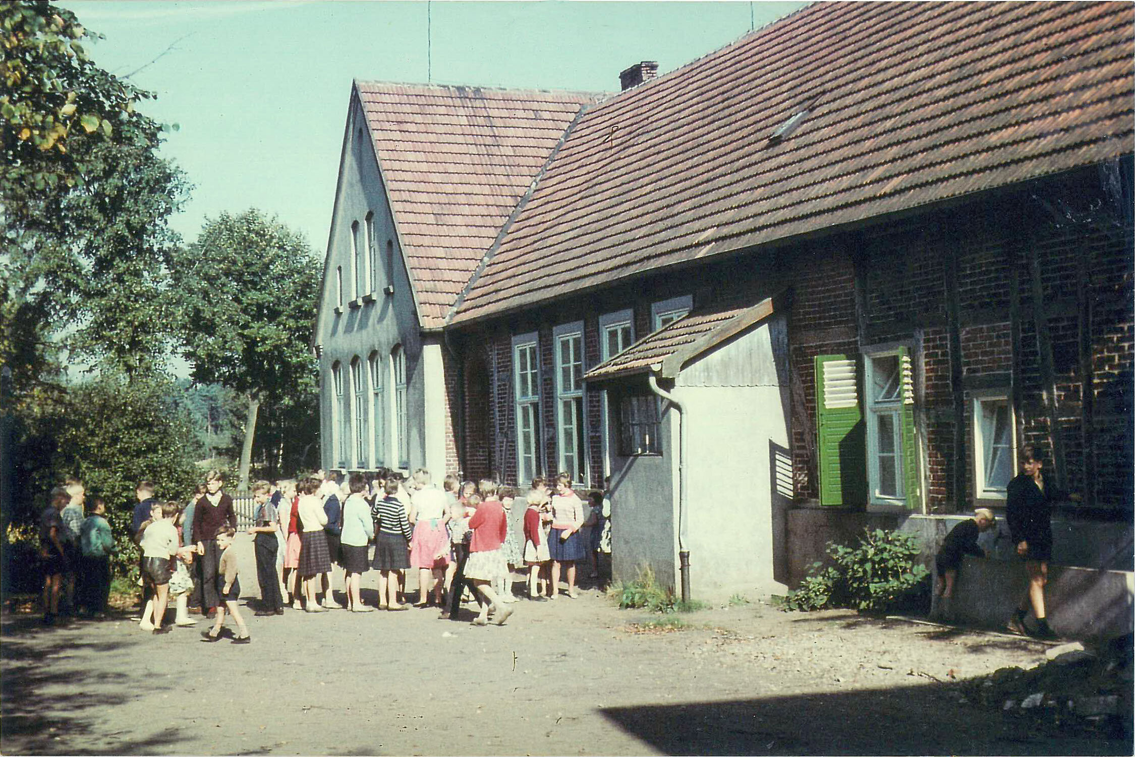 Eine Gruppe von Menschen versammelt sich vor einem Backsteingebäude mit rotem Dach und grüner Tür, umgeben von Bäumen und einem klaren blauen Himmel.