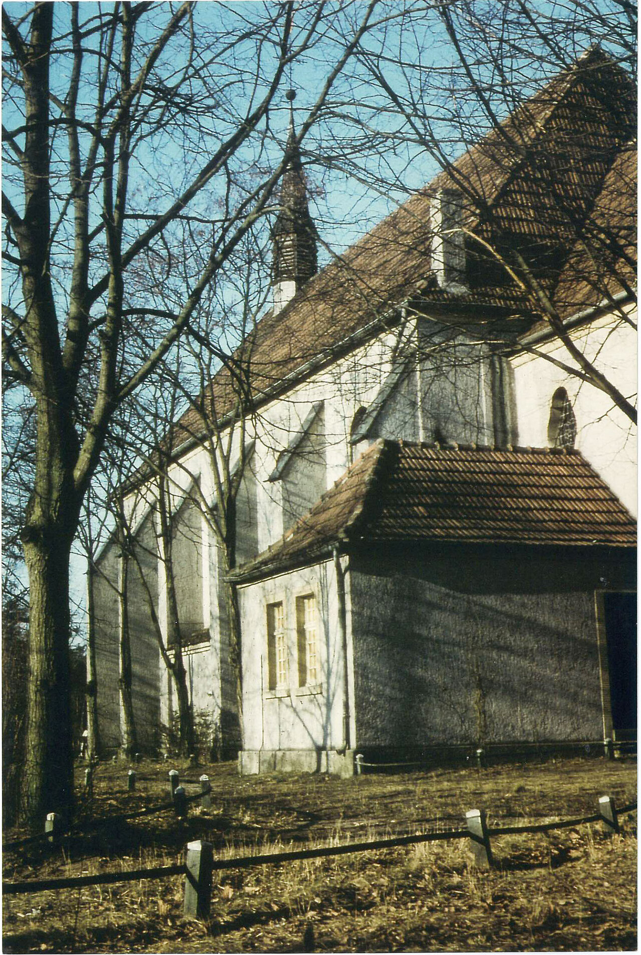 Eine weiße Kirche mit braunem Dach und Kirchturm steht in einer ruhigen Landschaft, umgeben von kahlen Bäumen und einem Holzzaun.