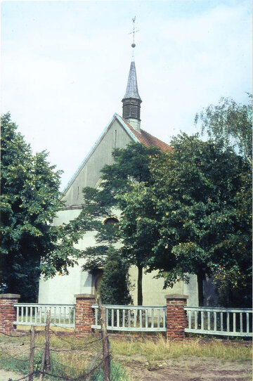 Inmitten von üppigem Grün und einem weißen Zaun steht eine weiße Kirche mit rotem Dach und einem hohen Kirchturm, der mit einem Kreuz geschmückt ist.