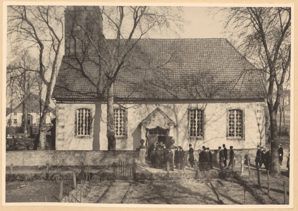 Schwarz-Weiß-Fotografie der Haustenbecker Kirche mit steilem Dach und Schornstein, umgeben von Bäumen und einer Gruppe von Menschen am Konfirmationstag, aufgenommen aus einem niedrigen Winkel.