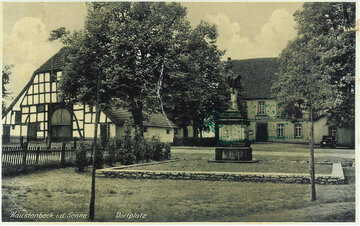 Eine Schwarz-Weiß-Fotografie eines Dorfplatzes in Haustenbeck, Deutschland, zeigt ein steinernes Denkmal, ein Fachwerkhaus und einen Baum, mit dem Text "Haustenbeck i.d. Senne" und "Dorfplatz" in der Ecke.