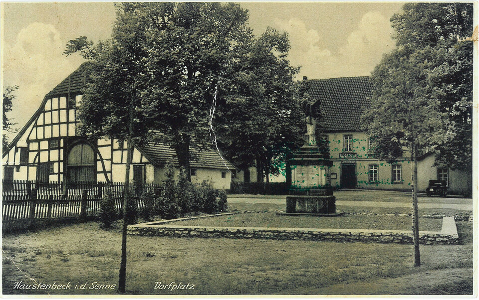 Eine Schwarz-Weiß-Fotografie eines Dorfplatzes in Haustenbeck, Deutschland, zeigt ein steinernes Denkmal, ein Fachwerkhaus und einen Baum, mit dem Text "Haustenbeck i.d. Senne" und "Dorfplatz" in der Ecke.