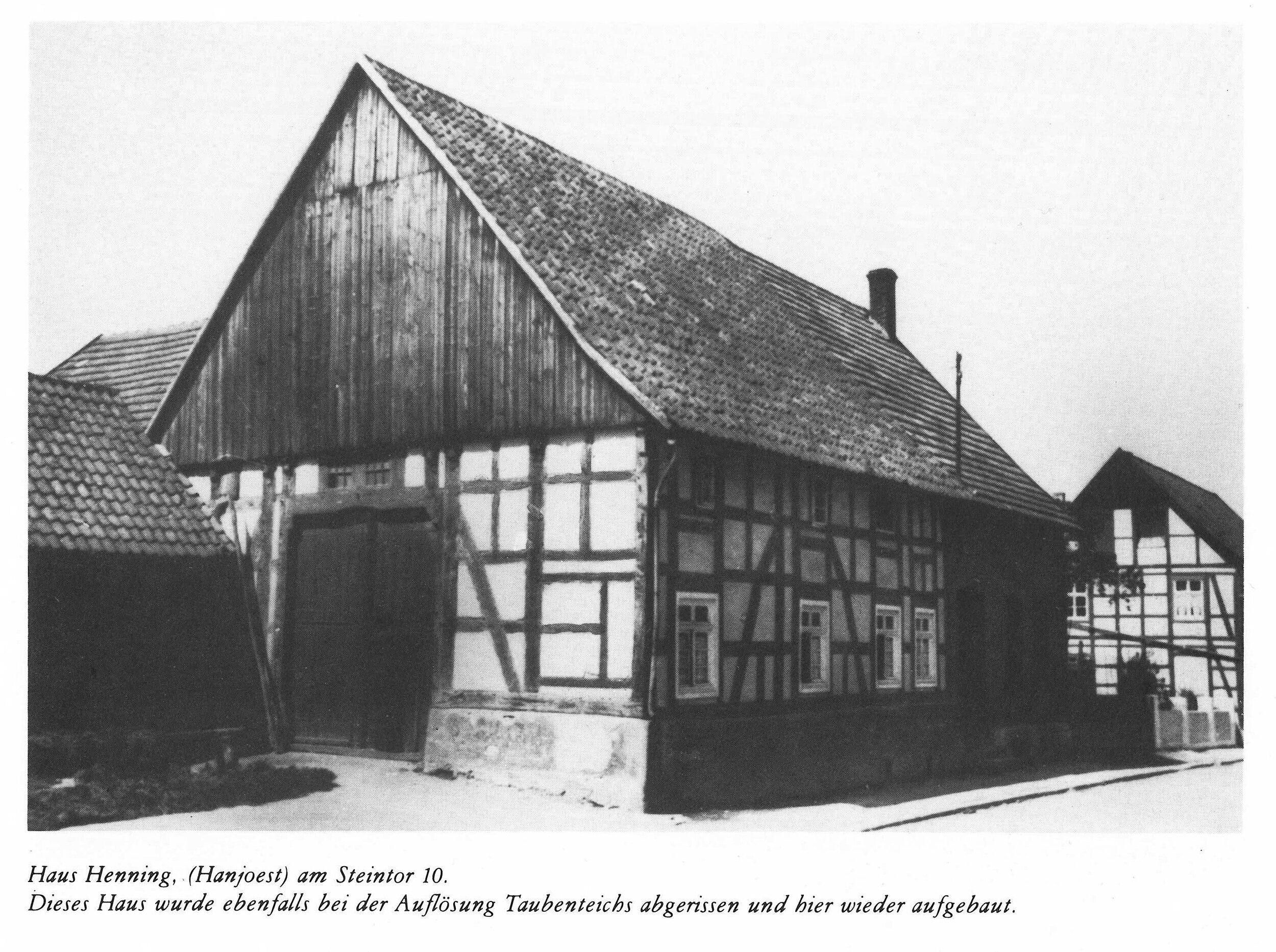Eine Schwarz-Weiß-Fotografie eines Fachwerkhauses mit steilem Dach und Schornstein, begleitet von einem kleineren Gebäude und einem Schild, das den Standort des Hauses angibt.