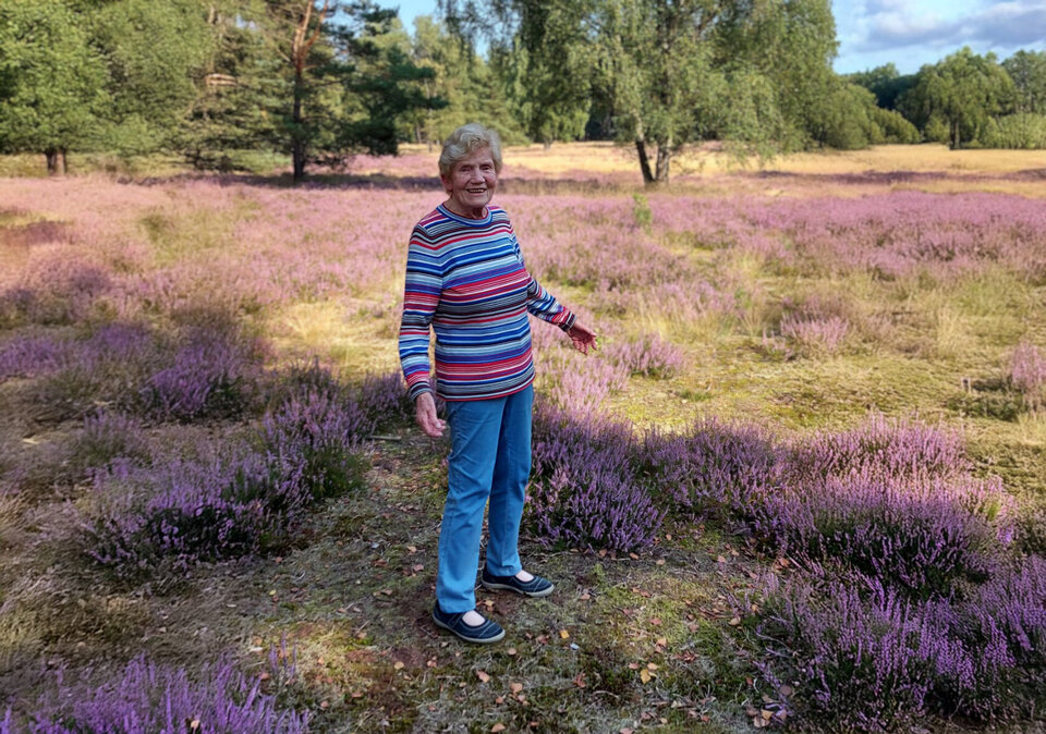 Zu sehen ist Frau Leonie Bolte in der alten Senne, inmitten eines Felds von Heidekräutern. Sie lacht fröhlich in die Kamera. Im Hintergrund sind Bäume zu sehen.