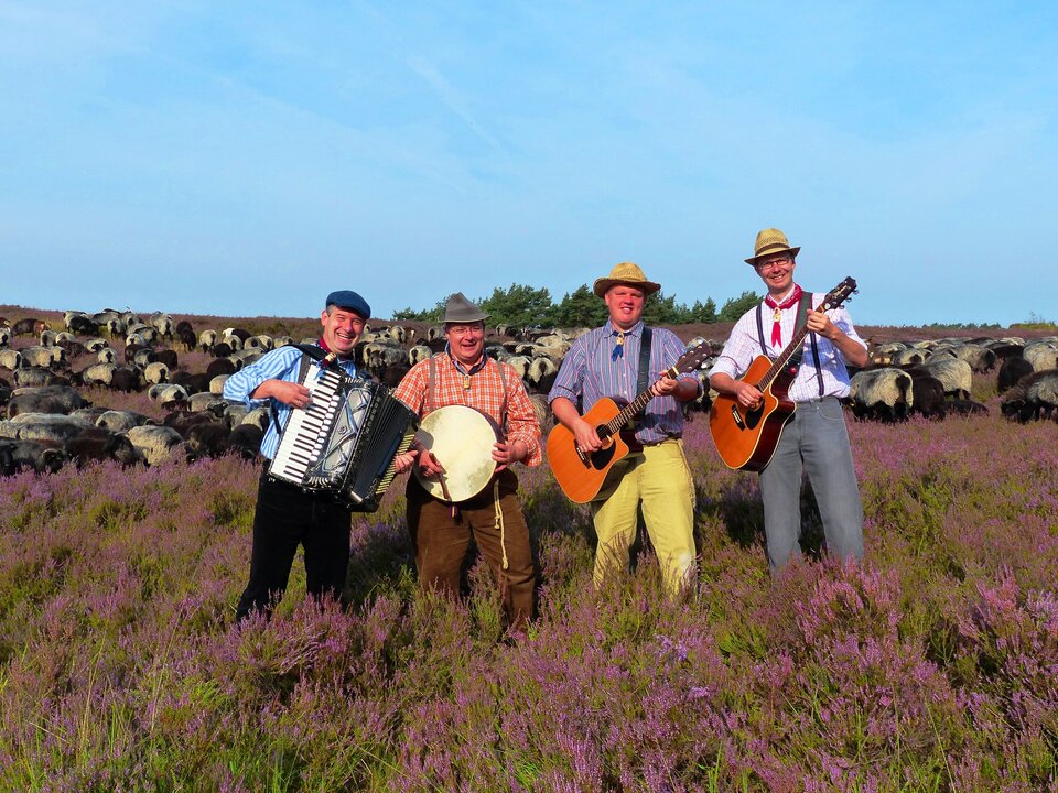 Zu sehen sind die Siene Puttkers, eine Band, in traditoneller Kleidung mit traditionellen Musikinstrumenten in der Senner Heide. Sie halten zum Beispiel ein Akkordeon, eine Gitarre und eine Trommel. Im Hintergrund sind blühende Heidekräuter und Heidschnucken zu sehen.
