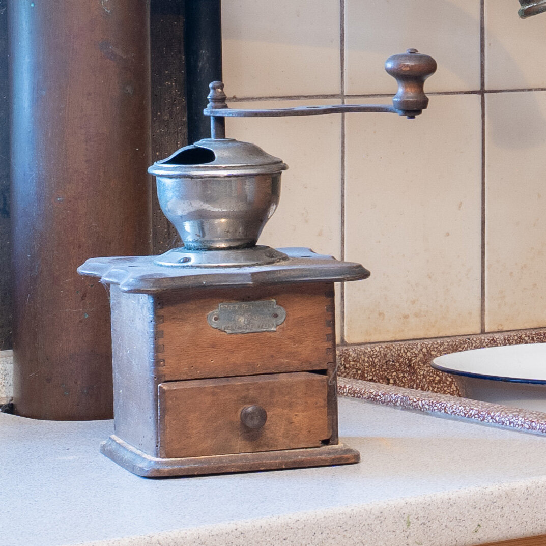 Zu sehen ist eine alte Kaffeemühle aus Holz und Metall. Sie steht auf einer alten weißen Arbeitsplatte mit weißen Kacheln im Hintergrund.