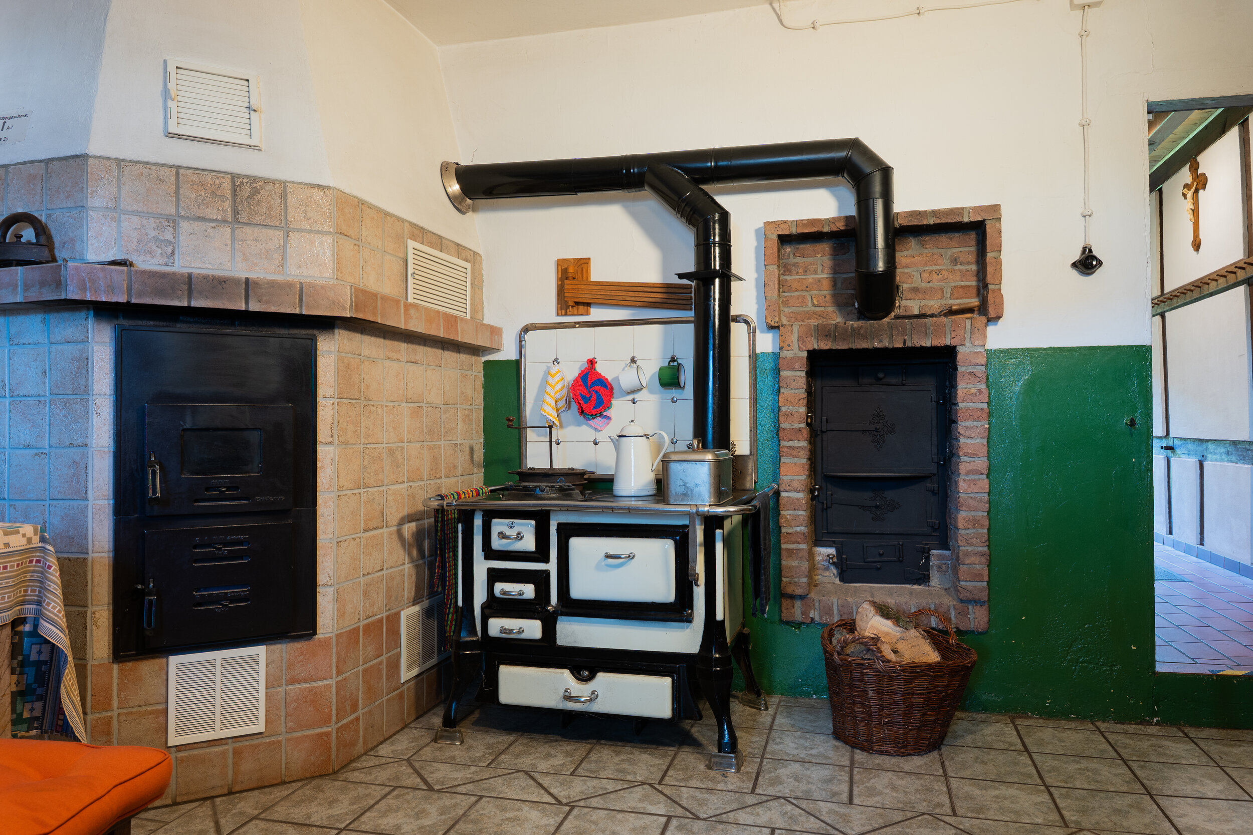 Zu sehen ist ein alter Backofen. Auf dem Backofen befindet sich eine Kanne. Daneben ein Feuerkorb mit Holz. Weiterhin in der Szene ist ein Kamin zu sehen.