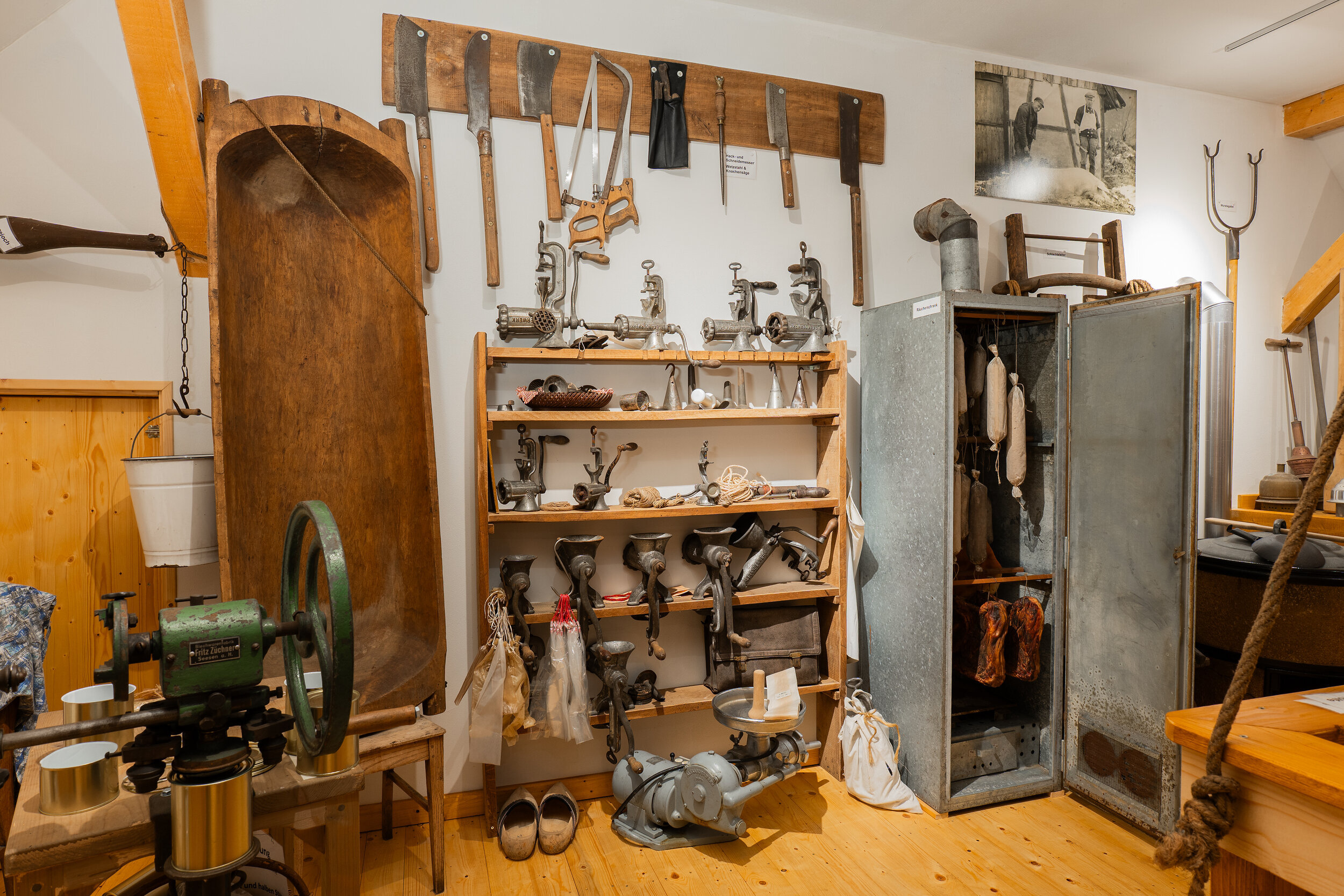 Auf dem Foto sind zahlreiche Instrumente und Hilfsmittel zur Fleischverarbeitung zu sehen. Einige Fleischwölfe, eine Blutwanne, ein Räucherschrank, Hackbeile und Sägen, sowie einiges mehr.