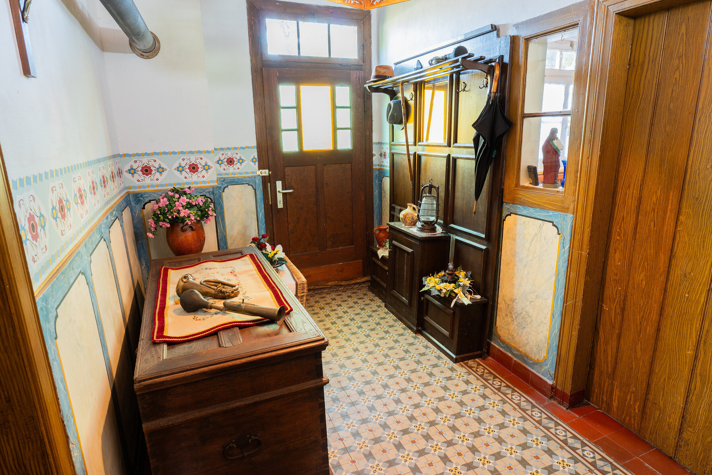 Das Bild zeigt einen Blick in einen alten, traditionellen Flur. Die Wände sind mit Holz verkleidet. Vor einer Tür steht eine große, alte Holztruhe. Die Wände sind zudem per Hand mit einem traditionellen Muster bemalt. An der Wand hängt zudem noch eine Garderobe mit Kleidung.