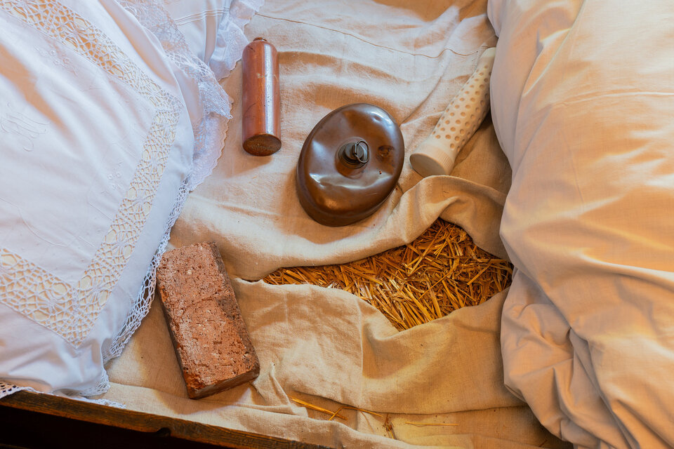 In einem alten Bett befinden sich ein weißes, spitzenbesetztes Kissen, eine braune Metallflasche, ein Backstein und eine Keramikflasche mit Sand.