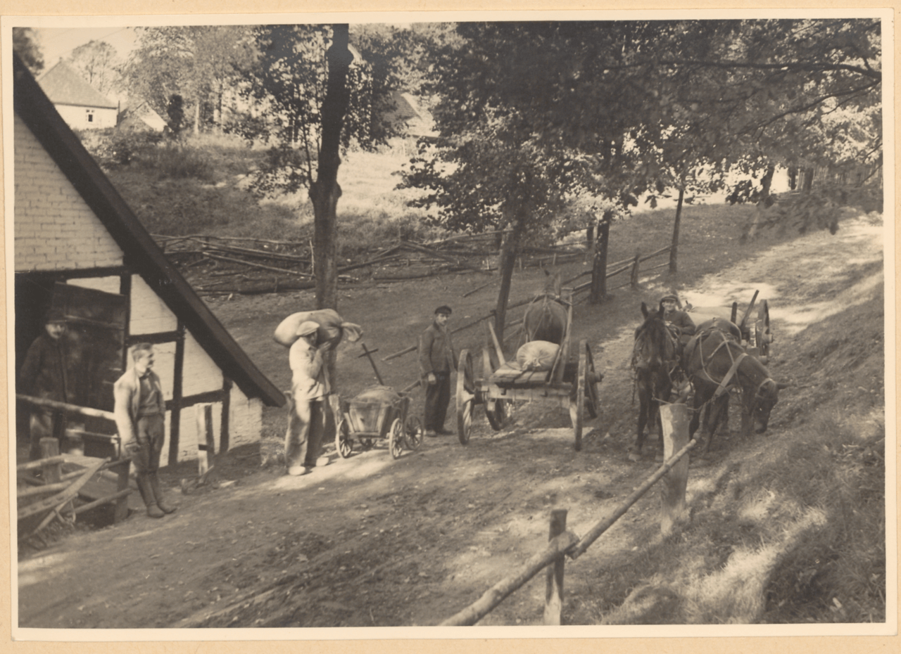 Eine Schwarz-Weiß-Fotografie zeigt eine ländliche Szene mit einem Pferdewagen, einem Mann mit einem Sack und einer Frau mit einem Korb, mit einem Holzzaun und Bäumen im Hintergrund, vor einer alten Mühle.