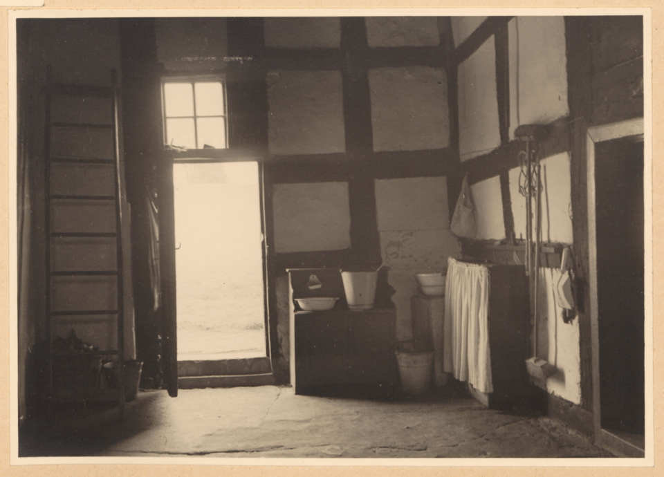 Eine Schwarz-Weiß-Fotografie einer rustikalen Deele mit Holztür, Fenster und Holzbank sowie Eimer, Schüssel und Besen auf einem Regal.