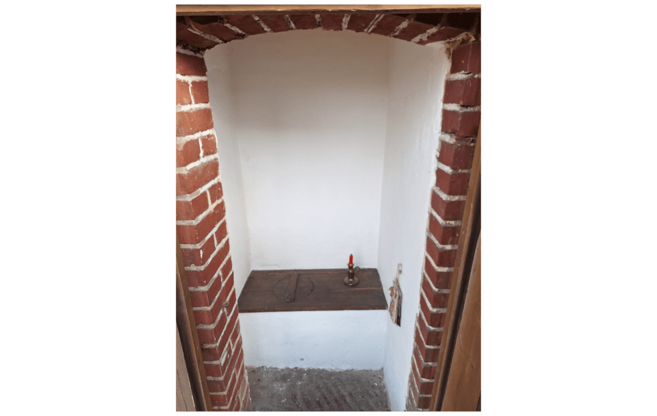 Ein Blick in ein altes Plumsklo. Zu sehen sind weiß gekalkte Wände, ein hölzerner Deckel und ein hölzerner Sitz. Auf der Ablage steht ein Kerzenhalter aus Metall mit einer Kerze.