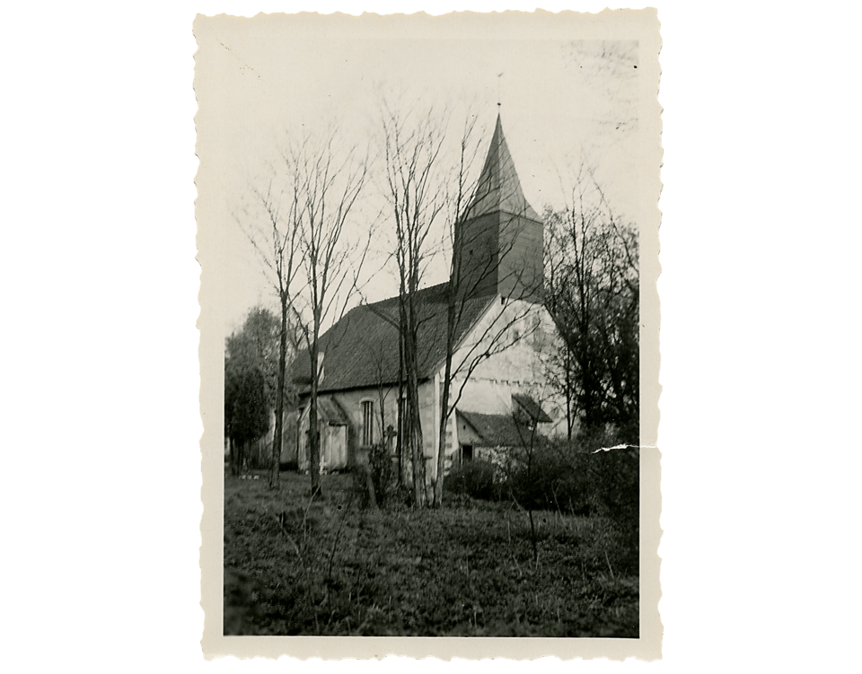 Die Haustenbecker Kirche in weiß, mit Kirchturm und Kreuz steht in einer ländlichen Umgebung, umgeben von Bäumen und Sträuchern, mit einer Schwarz-Weiß-Fotografie.