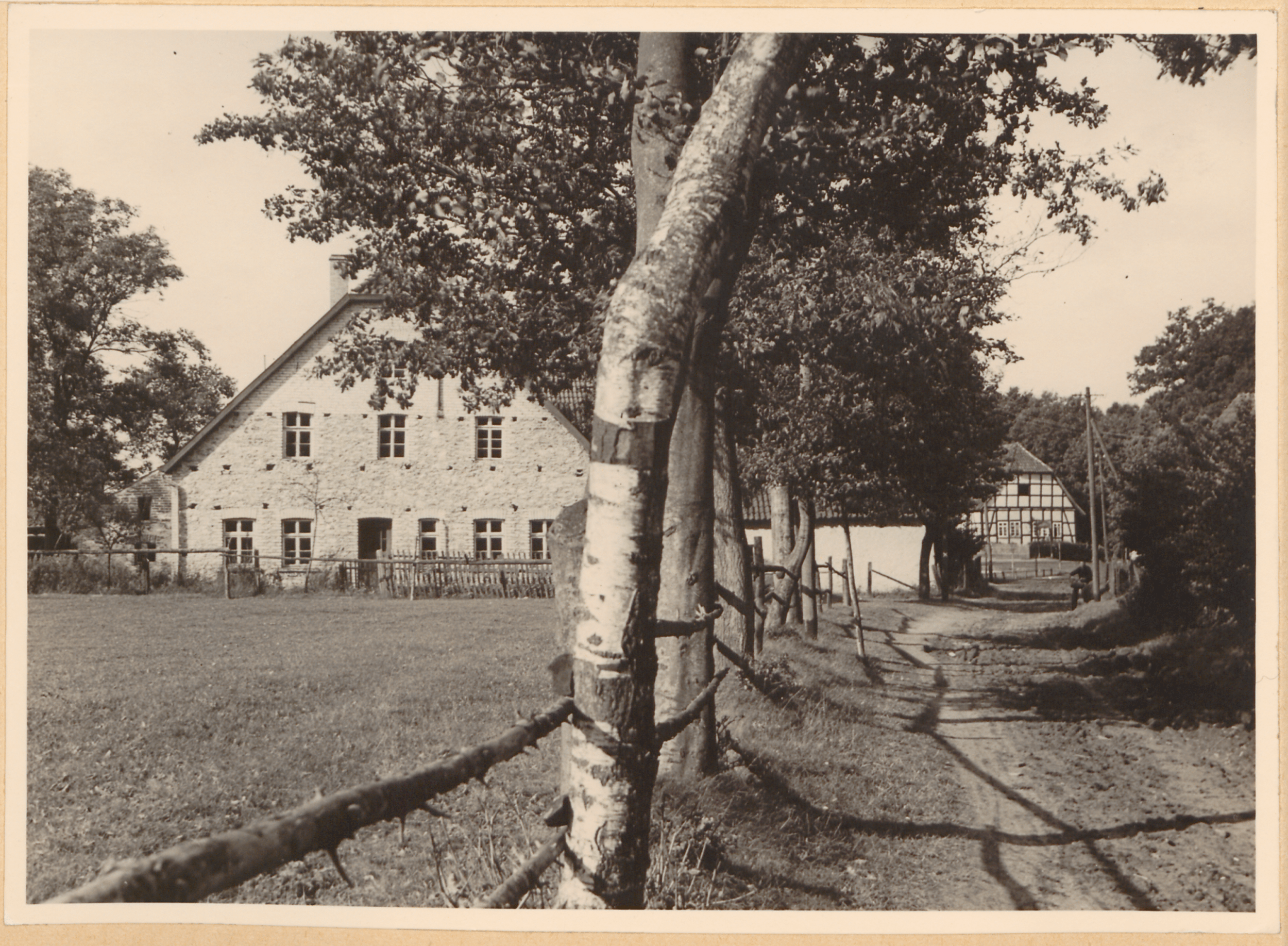 Eine Schwarz-Weiß-Fotografie zeigt ein großes Steinhaus mit steilem Dach, umgeben von einem Holzzaun und einem Feldweg, mit Bäumen und einem kleineren Gebäude im Hintergrund.