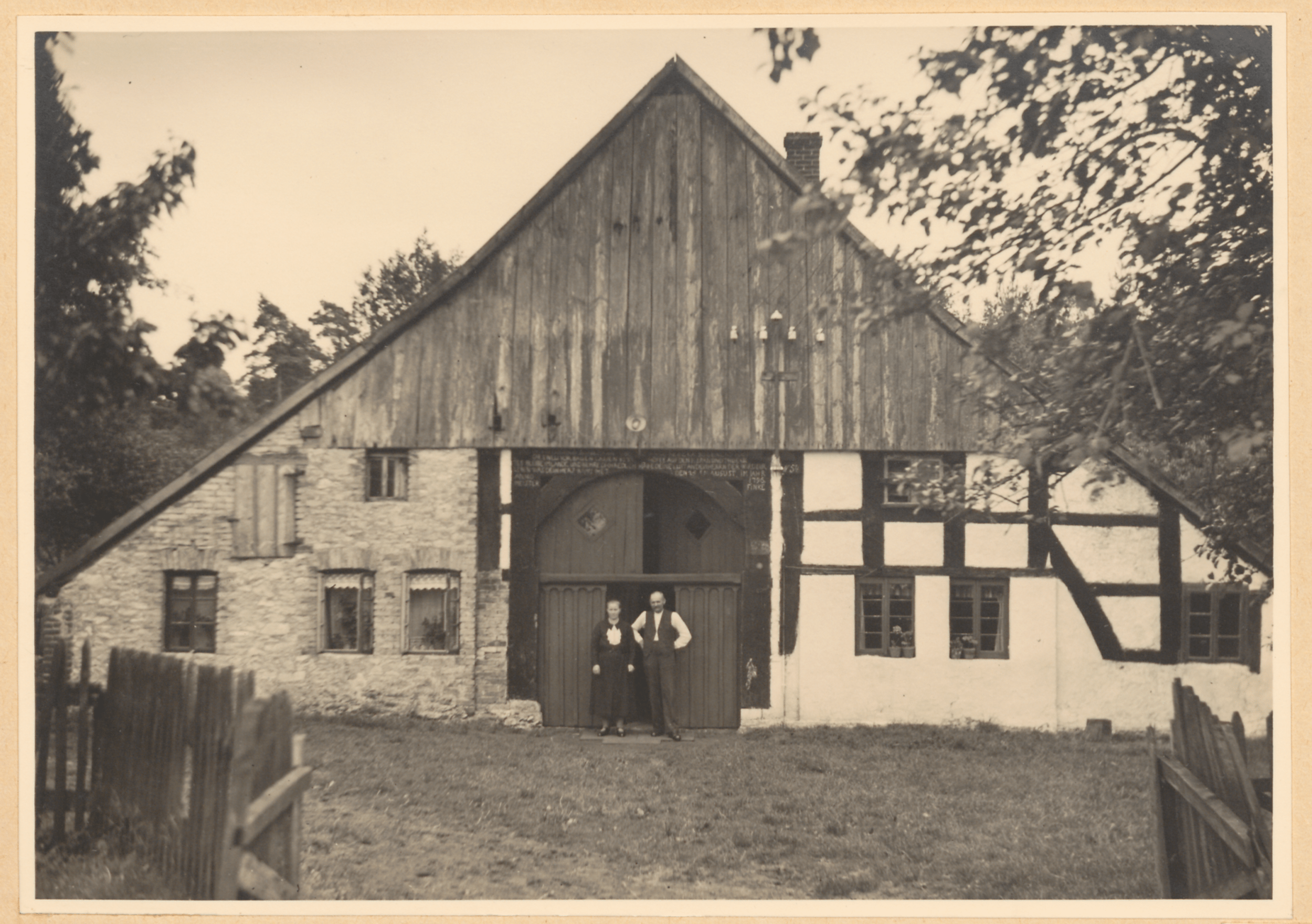 Eine Schwarz-Weiß-Fotografie einer großen Holzscheune mit steilem Dach, weißen Wänden und einer Holztür, umgeben von einem Holzzaun und Bäumen.
