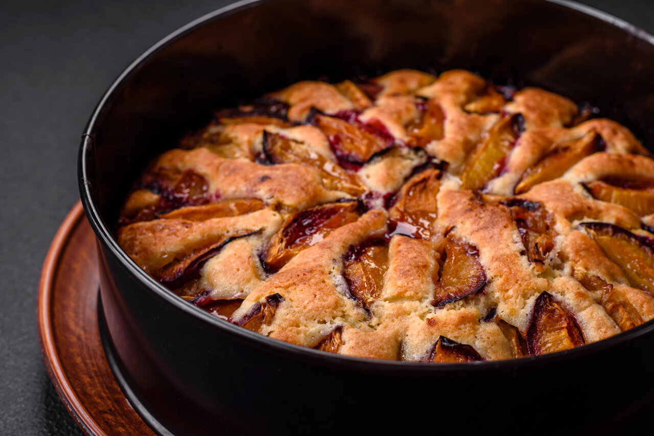 Ein goldbrauner Obstkuchen, belegt mit Zwetschgen, steht in einer schwarzen Auflaufform auf einem Holzteller und ist bereit zum Servieren.