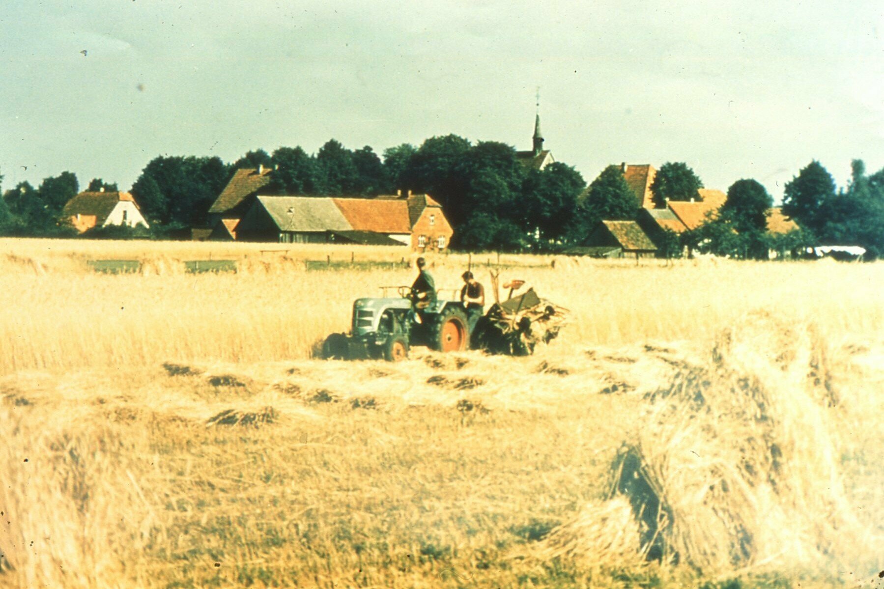 Ein blauer Traktor zieht einen großen Pflug durch ein goldenes Weizenfeld, mit einem kleinen Dorf und einer Kirche in der Ferne, unter einem klaren blauen Himmel.