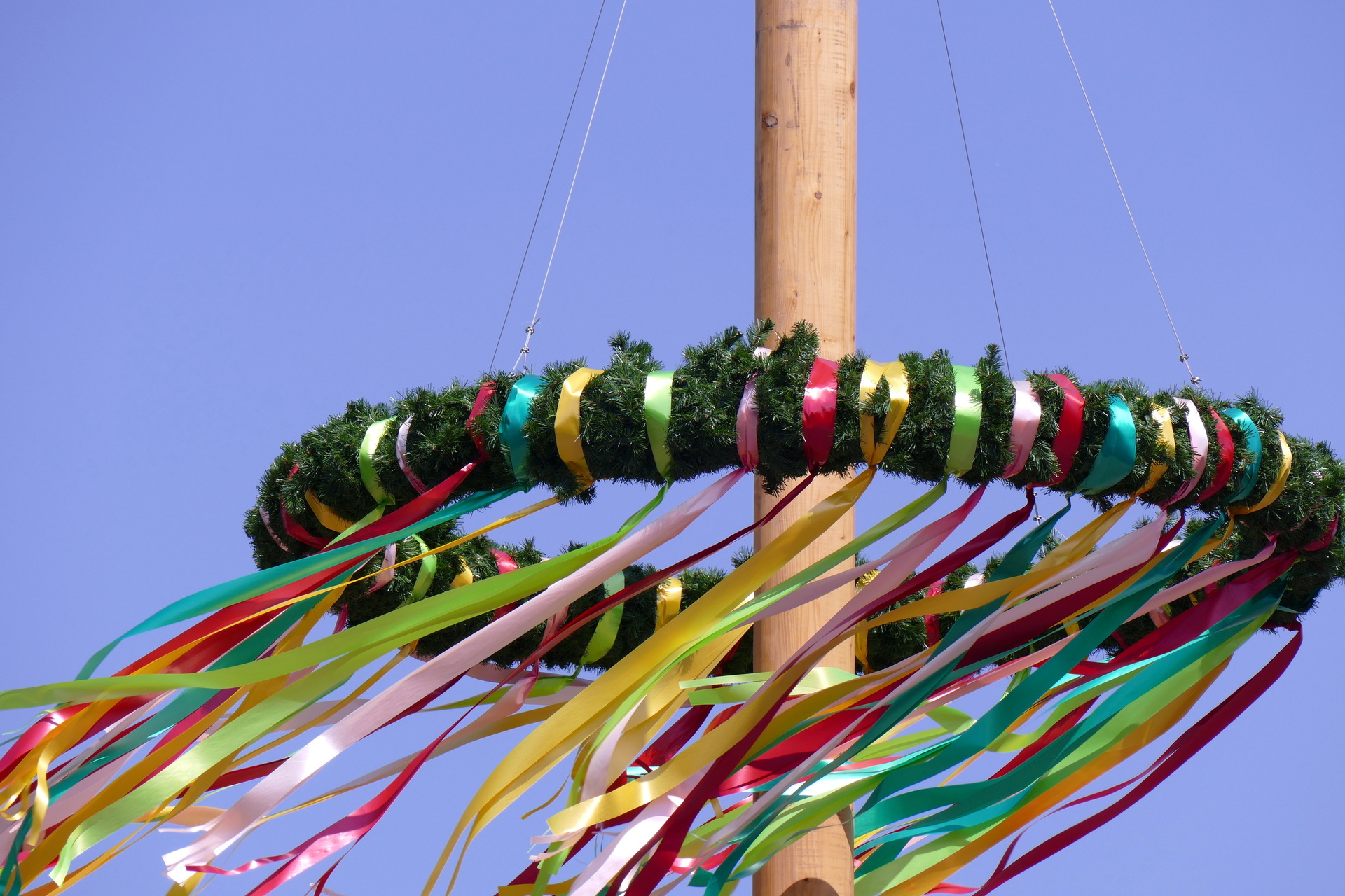 Eine Holzstange trägt einen Maibaumkranz für ein Schützenfest. An dem Kranz hängen viele bunte lange Bänder, die im Wind wehen.