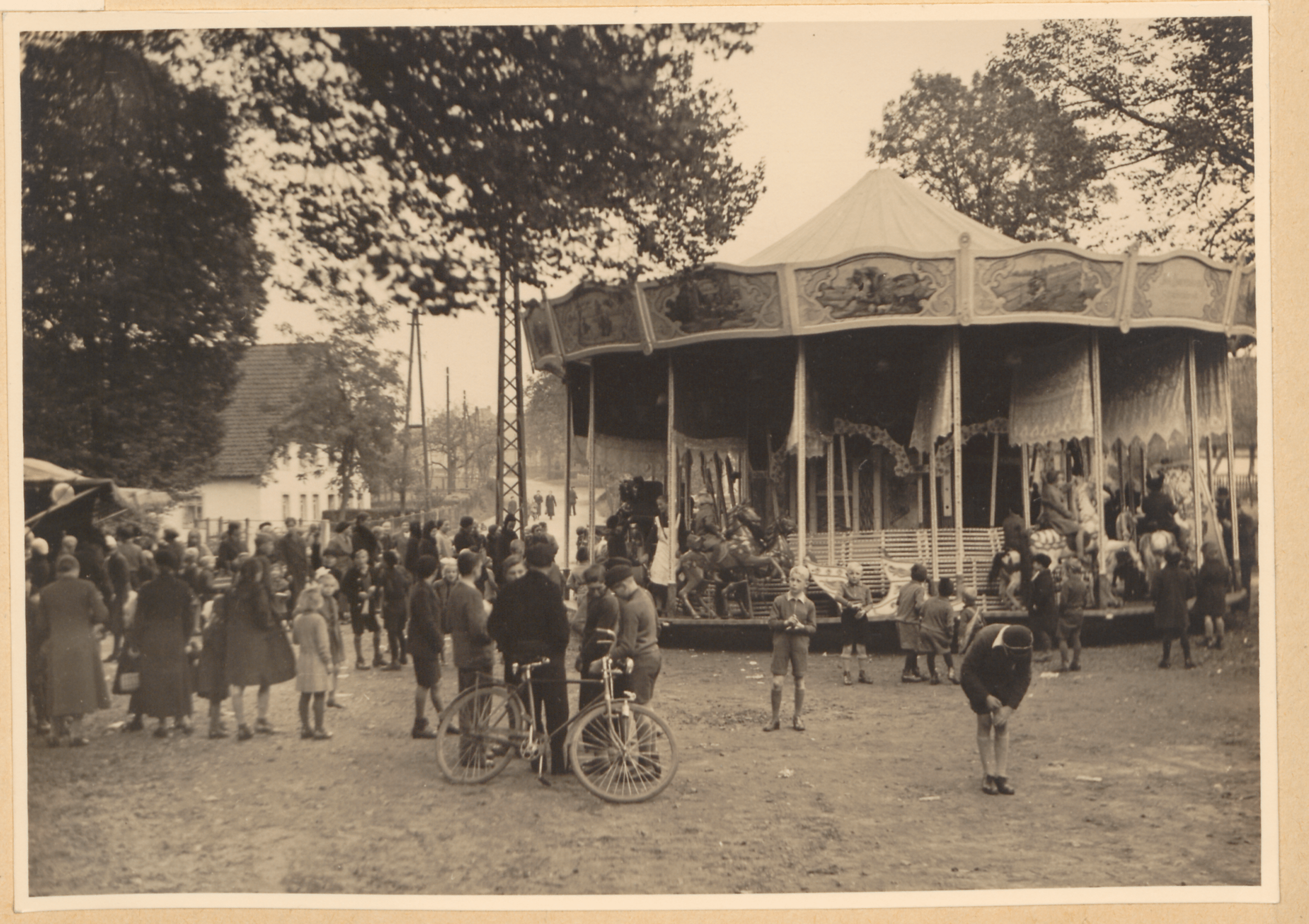 Ein belebtes Karussell mit Pferden und Menschen steht in einem Park, umgeben von Bäumen und einem weißen Gebäude, mit einer Schwarz-Weiß-Fotografie im Vordergrund.